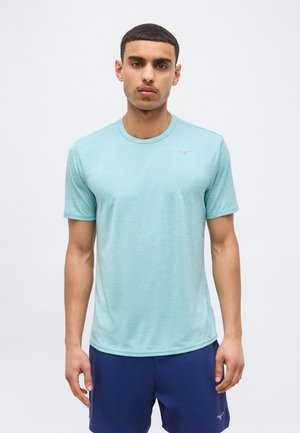 Άνδρας που στέκεται φορώντας ανοιχτό μπλε αθλητικό πουκάμισο με κοντά μανίκια και σκούρα μπλε σορτς, μπροστά σε απλό φωτεινό φόντο.