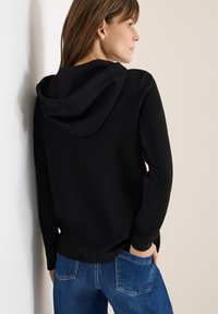 Černá mikina s uvolněným střihem, s kapucí na šňůrku, dlouhými rukávy a jemnou strukturou. V kombinaci s modrými džíny.
