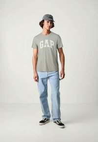 Γκρι t-shirt με το λογότυπο "GAP" σε λευκό και κόκκινο, συνδυασμένο με ανοιχτό μπλε τζιν και μαύρα sneakers. Το μοντέλο φορά επίσης ένα γκρι καπέλο.