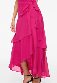 Femme portant une jupe midi en mousseline fuchsia à volants, avec un ourlet asymétrique et une taille nouée, associée à des talons noirs à brides à la cheville.