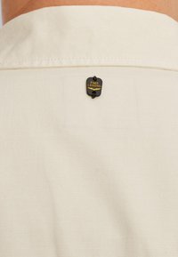 Tissu de chemise beige clair présentant un badge noir avec le texte "PME Legend", attaché au cou par une petite boucle noire.