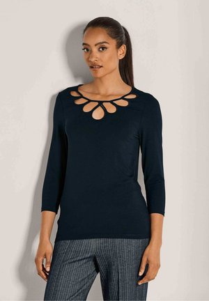 Vrouw met lang, donker haar in een paardenstaart, gekleed in een zwart longsleeve top met druppelvormige uitsparingen rond de halslijn en grijs gestreepte broek.