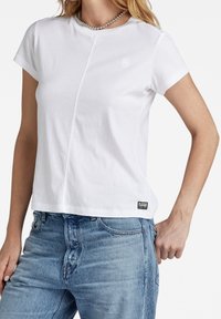 T-shirt blanc à manches courtes en coton, avec un détail de couture sur le devant et un petit logo sur la poitrine gauche. Associé à un jean bleu.