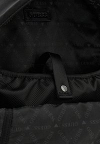 Intérieur noir d'un sac avec un patch logo GUESS, texture lisse et une fine sangle avec un bouton-pression. Motif de marque subtil partout.