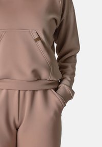 Světlobéžová mikina s velkou přední kapsou, žebrovanými manžetami a odpovídajícími zúženými kalhotami. Hladká textura, minimalistický design, bez vzorů.