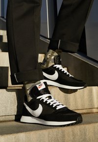 Baskets Nike noires et blanches avec des lacets blancs, portées avec des chaussettes camouflage et un pantalon noir à revers sur des marches en béton à l'extérieur.
