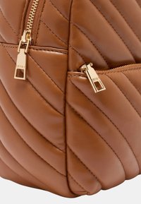 Zaino in pelle marrone con design trapuntato, caratterizzato da cuciture diagonali e dettagli in metallo color oro sulle cerniere. Comprende anche una tasca aggiuntiva con cerniera.