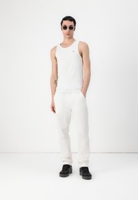 Fehér ujjatlan felső és laza szabású világos nadrág, fekete sportcipővel párosítva. A modellen sötét napszemüveg van. Minimalista design fehér háttérben.