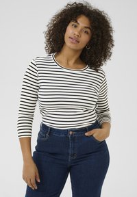 Striped long-sleeve top in white and black, with a round neckline. Paired with high-waisted dark denim jeans. Curly hair, hands resting on hips.