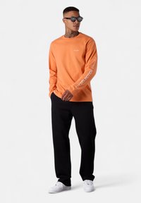 Langärmliges oranges Baumwollhemd mit weißem Text am Ärmel, kombiniert mit schwarzen Hosen und weißen Sneakern, getragen von einer Person mit Sonnenbrille.