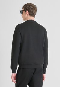 Felpa nera con scollo rotondo, maniche lunghe e texture liscia; indossata con pantaloni neri, che mostrano una vestibilità aderente e un design minimale.