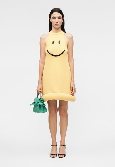 Γυναίκα στέκεται φορώντας κίτρινο φόρεμα χωρίς μανίκια με μαύρο χαμογελαστό πρόσωπο, κρατά μικρή πράσινη τσάντα και φορά μαύρα ανοιχτά παπούτσια με τακούνι.