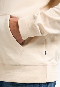 Sudadera beige en suave mezcla de algodón, con bolsillo tipo canguro, puños y dobladillo acanalados, y una pequeña etiqueta de logo en el lado inferior.