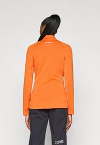 Oransje langermet treningsgenser med teksturert ribbing, høy krage og logo bak i nakken. Kombinert med mørkfargede bukser.