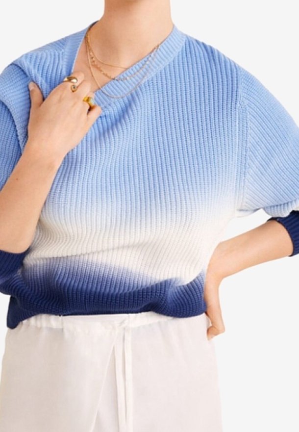 Femme portant un pull en tricot ombré bleu vers blanc, pantalon blanc, bagues dorées et colliers dorés superposés, main touchant la clavicule.