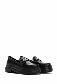 Mocasines de piel negra con suela gruesa, que presentan acentos de hardware plateado y una textura suave. Diseño de perfil bajo con punta redondeada.