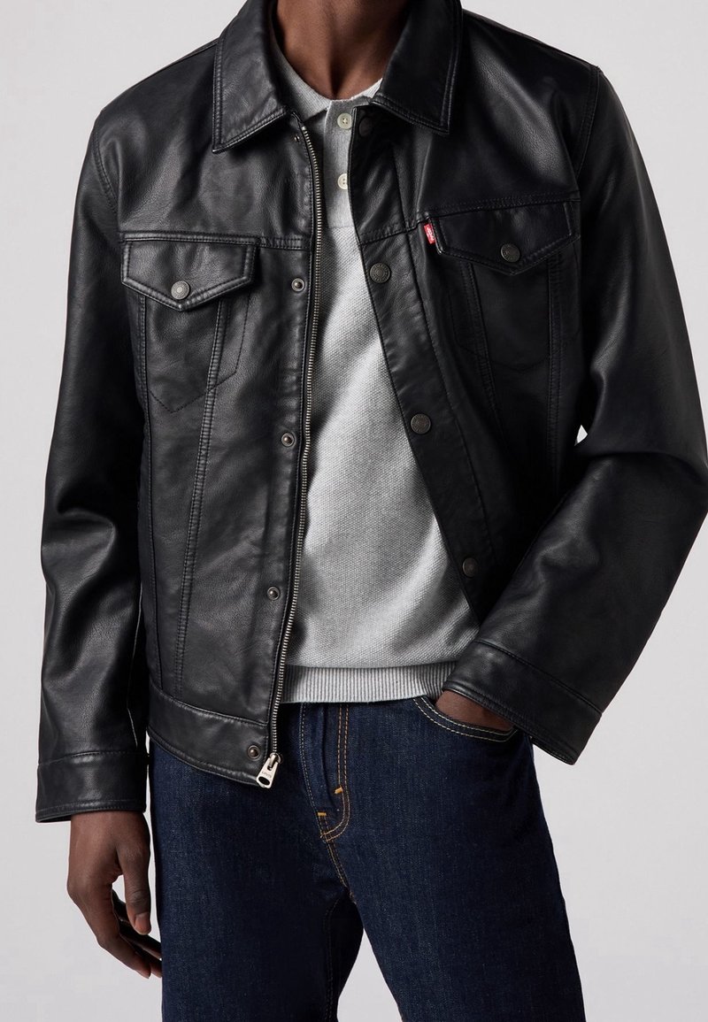 Giacca in pelle nera con tasche sul petto con bottoni, colletto classico e hardware argentato. Abbinata a un maglione grigio e jeans blu.