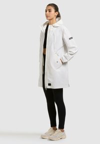 Kvinde stående iført en lang hvid hættetrøje regnjakke over sorte leggings og beige sneakers, hænder i jakkelommerne, vendt mod venstre.