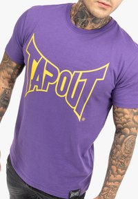 Lila pamut póló, nagy sárga "TAPOUT" logóval az elején. Rövid ujjú és kerek nyakú kialakítás. Címke látható a szegélyen.