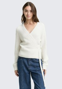 Bela ovita jopica, izdelana iz mehkega, puhastega materiala, z globokim V-izrezom, širokimi manšetami in veznim pasom v pasu. Uporabljena s modrimi jeans hlačami.