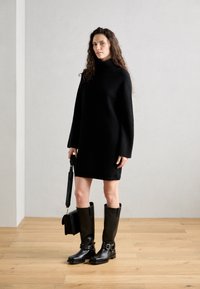 Čierne rebrované pletené šaty s vysokým golierom, spárované s čiernymi koženými čižmami po kolená s prackami. Modelka drží malú čiernu tašku.