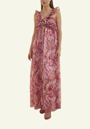 Frau trägt ärmelloses, bodenlanges, rosa- und lila-blumiges Kleid mit Rüschen, tiefem V-Ausschnitt und offenen Sandalen, steht vor einfachem Hintergrund.