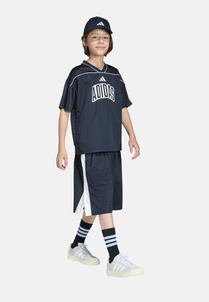 Niño con gorra negra de Adidas, camiseta negra de Adidas holgada, pantalones cortos negros con rayas blancas, calcetines negros con rayas blancas y zapatillas blancas de Adidas.