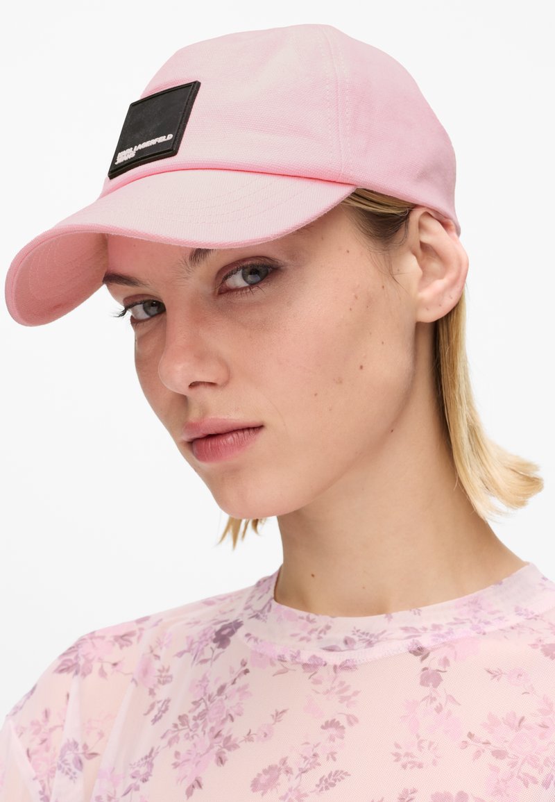 Κοντινό πλάνο ενός ατόμου που φοράει ροζ ανοιχτό καπέλο με μαύρο σημαιάκι και διάφανο ροζ τοπ με floral σχέδιο, κοιτάζοντας ελαφρώς προς τα αριστερά.