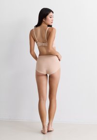 Jonge vrouw staat blootsvoets met haar rug naar de camera, gekleed in naadloze huidkleurige ondergoed en beha voor een effen witte achtergrond.