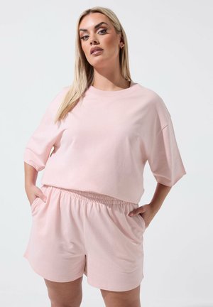 Kobieta o długich blond włosach, ubrana w jasnoróżową, oversize'ową bluzkę z krótkim rękawem oraz pasujące krótkie spodenki z elastycznym pasem, z rękami w kieszeniach.