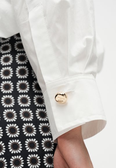 Main portant un poignet de chemise blanc fixé par un bouton de manchette rond en or gravé "Sandro", sur un tissu à motif floral noir et blanc.