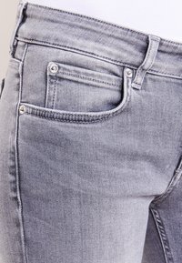 Jeans de mezclilla gris claro con una vista en primer plano, que presentan un bolsillo, herrajes plateados y un detalle de textura sutil en la tela.