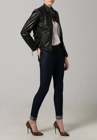 Giubbotto di pelle nera con colletto alto, abbinato a una camicia bianca, jeans di denim scuro e tacchi alti con stampa leopardata.