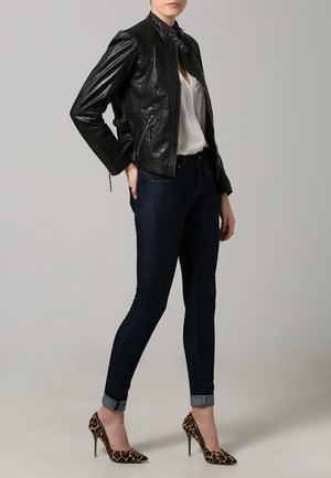 Veste en cuir noir avec col montant, associée à une blouse blanche, un jean en denim foncé et des escarpins à imprimé léopard.