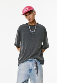 Dunkelgraues, übergroßes T-Shirt mit fransigen Kanten und strukturiertem Design. Kombiniert mit hellblauen Jeans und einer pinken Mütze.