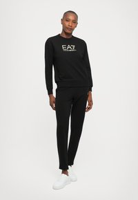 Egy nő fekete EA7 Emporio Armani pulóverben és ahhoz illő nadrágban, fehér sportcipővel, magabiztosan áll egy sima fehér háttér előtt.