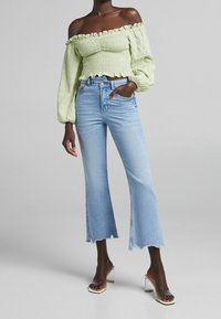 Donna che indossa un top smock off-shoulder verde chiaro, jeans cropped azzurri con orlo sfilacciato e sandali con tacco trasparente, in piedi contro uno sfondo grigio.