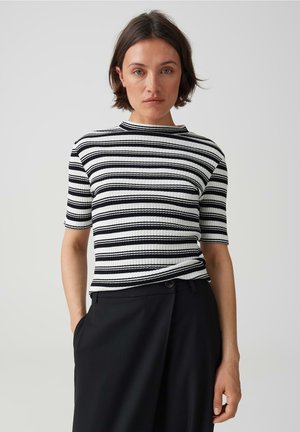 Donna con capelli corti e scuri che indossa una maglietta a righe bianche e nere a maniche corte a coste e pantaloni neri a vita alta, in piedi davanti a uno sfondo semplice.