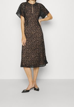 Kobieta ubrana w czarną koronkową sukienkę do połowy długości z krótkimi rękawami i cielistą podszewką, w zestawieniu z czarnymi szpiczastymi sandałkami na obcasie typu slingback z srebrnymi kokardkami.