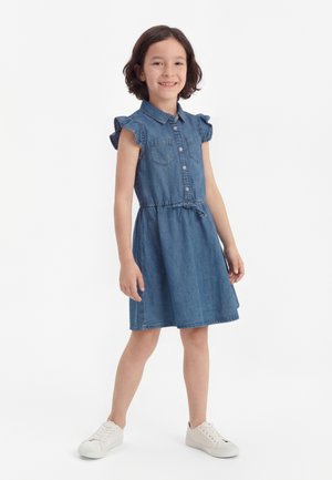 Nasmejana deklica stoji, oblečena v modro denim obleko brez rokavov z gumbi spredaj in belimi supergami na enobarvnem belem ozadju.