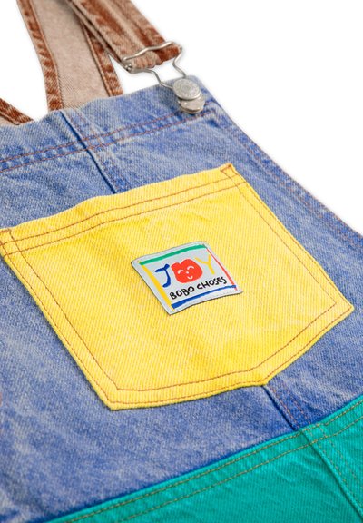 Denim overalls featuring a blue upper portion, yellow back pocket with a logo tag, and a green lower section. Adjustable brown straps included.