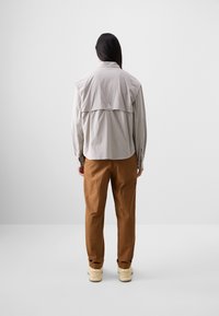Light gray long-sleeve shirt with a back yoke and triangular logo, paired with brown pants and beige footwear. Simple, functional design.