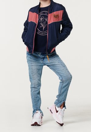 Niño con chaqueta azul marino y rosa, camiseta gráfica azul marino, jeans azules con dobladillo partido y zapatillas blancas con rosa, de pie con las manos en los bolsillos.