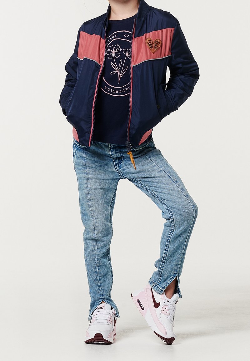 Bambino con giacca blu navy e rosa, t-shirt blu navy con grafica, jeans blu con orlo a spacco e scarpe bianche e rosa, in piedi con le mani nelle tasche.