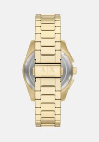 Zlatni metalni ručni sat s heksagonalnim kućištem, povezanim narukvicama i ugraviranim inicijalima branda na kopči. Reflektivni srebrni brojčanik je vidljiv.