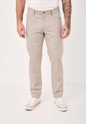 Mann trägt beige Slim-Fit-Hosen und weiße Turnschuhe, steht vor einem schlichten weißen Hintergrund, Tätowierungen an den Armen sichtbar.