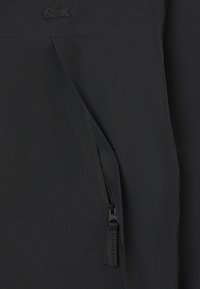 Giacca a vento nera con tasca con zip, texture liscia e logo Lacoste, dal design moderno e snello.
