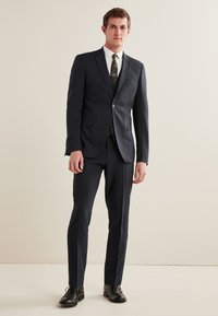 Marineblauer Anzug mit taillierter Passform, einreihiger Jacke, zwei Knöpfen, kontrastierendem weißen Hemd, gemusterter Krawatte und schwarzen Lederschuhen.