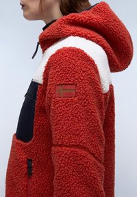 Red fleece jacket with a hood, white upper panel, and black pocket. Features textured fabric and a patch with a flag on the left sleeve.