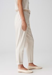 Pantalons beiges, fuselés avec des ourlets retroussés, présentant une texture de tissu lisse et des poches latérales. Accompagnés de ballerines simples de couleur crème.